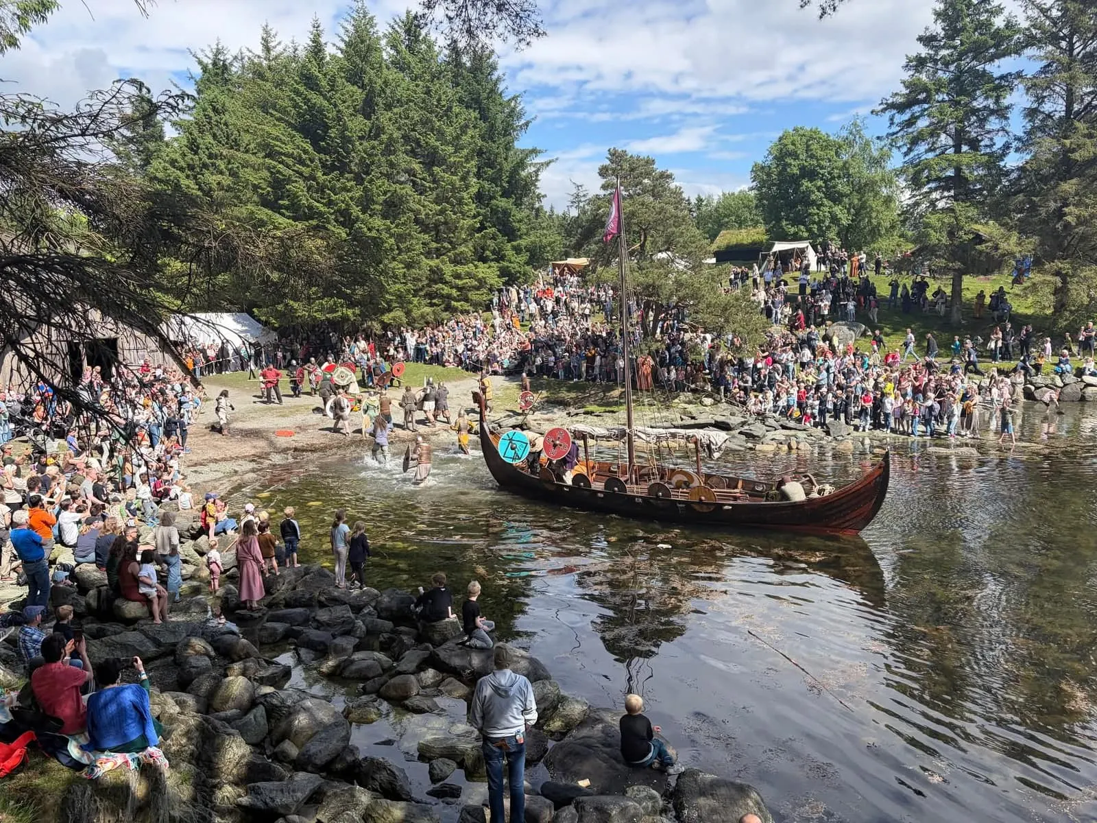 A replica Viking ship with painted shields along the side lies at the shore while armed Vikings splash through the shallows toward the defenders on the beach by a wooden boathouse, with hundreds of spectators on the rocks and the slope behind