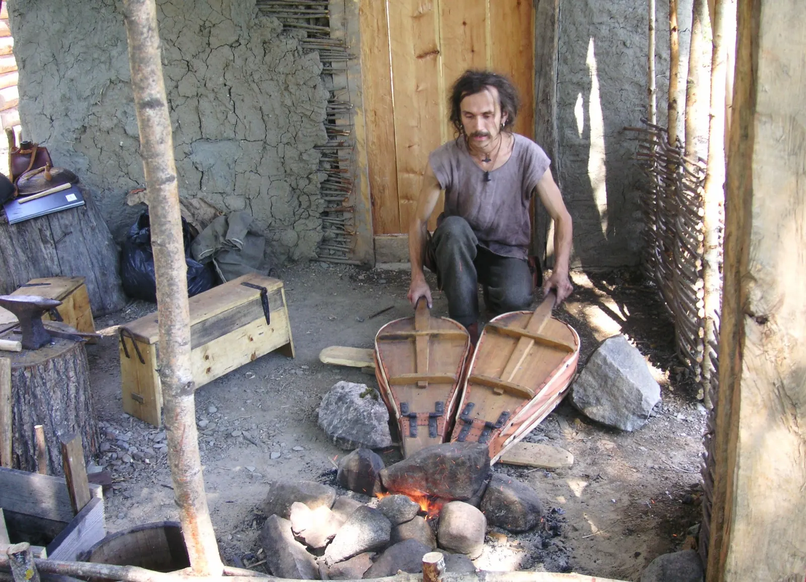 A costumed craftsman in a grey tunic working at an open hearth inside a wattle and daub workshop in the reconstructed Birka village