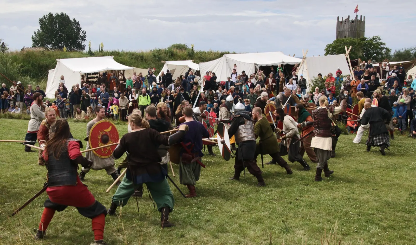 Dozens of reenactors with spears and round shields charging across a field during the battle show, market tents, spectators and a wooden watchtower behind