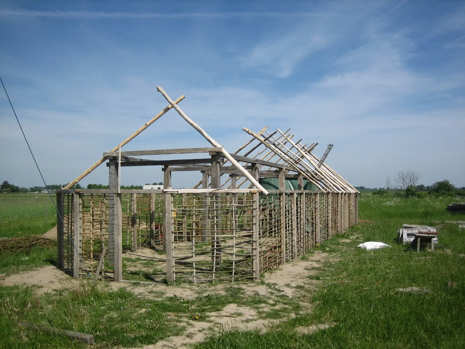 A Viking Age longhouse under construction at VikingaTider, its roof timbers and woven wattle walls standing open against a flat Skåne sky
