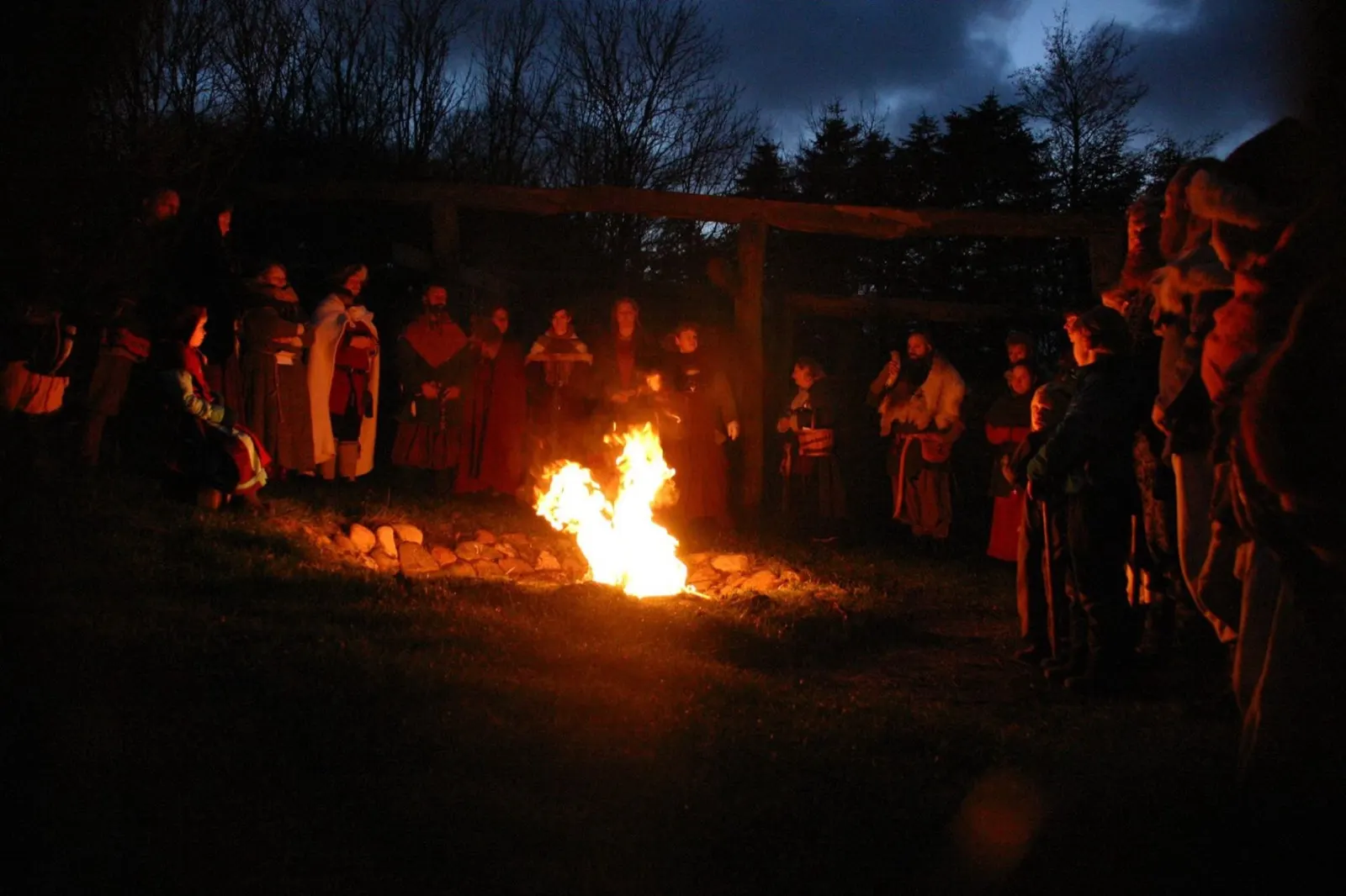 People in Viking dress stand in a wide circle around a fire on the grass at dusk, under a timber frame, with bare trees and a dark blue sky behind