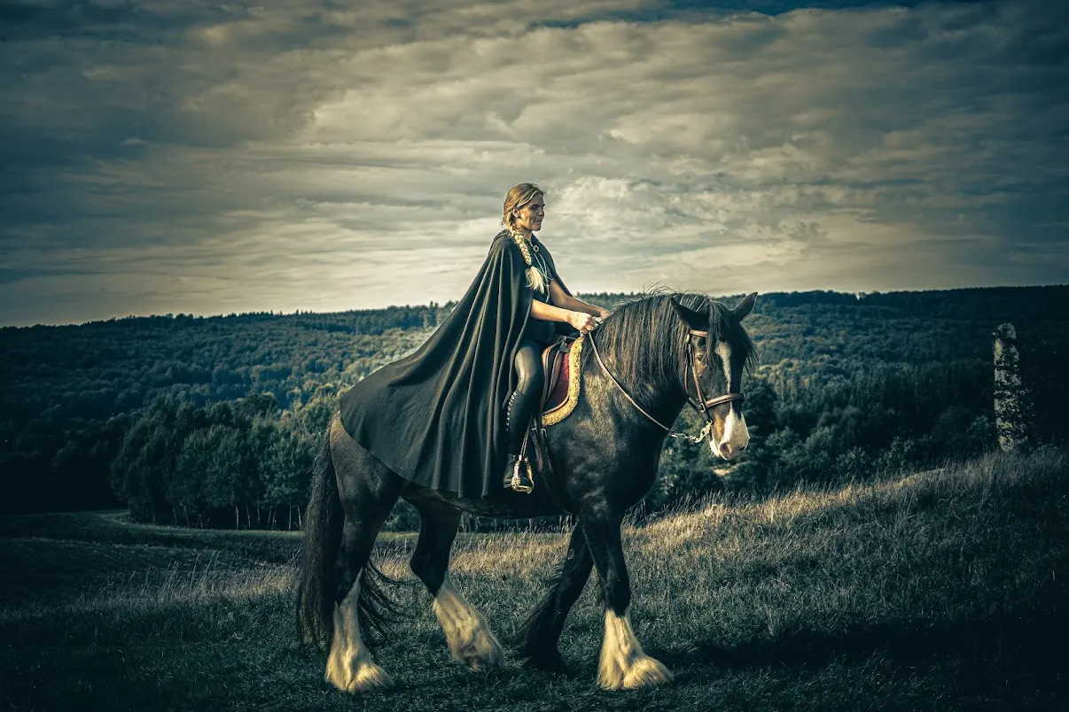 A rider in a long dark wool cloak on a black horse with white feathered legs, crossing a grassy hilltop at Gudahagen with wooded ridges behind under a heavy sky