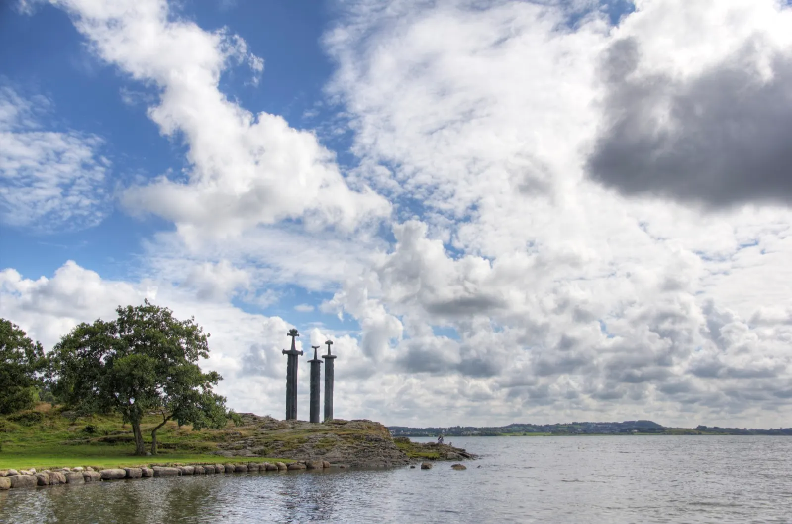 The three bronze swords of the Sverd i fjell monument standing on a rocky point by the fjord under a dramatic cloudy sky