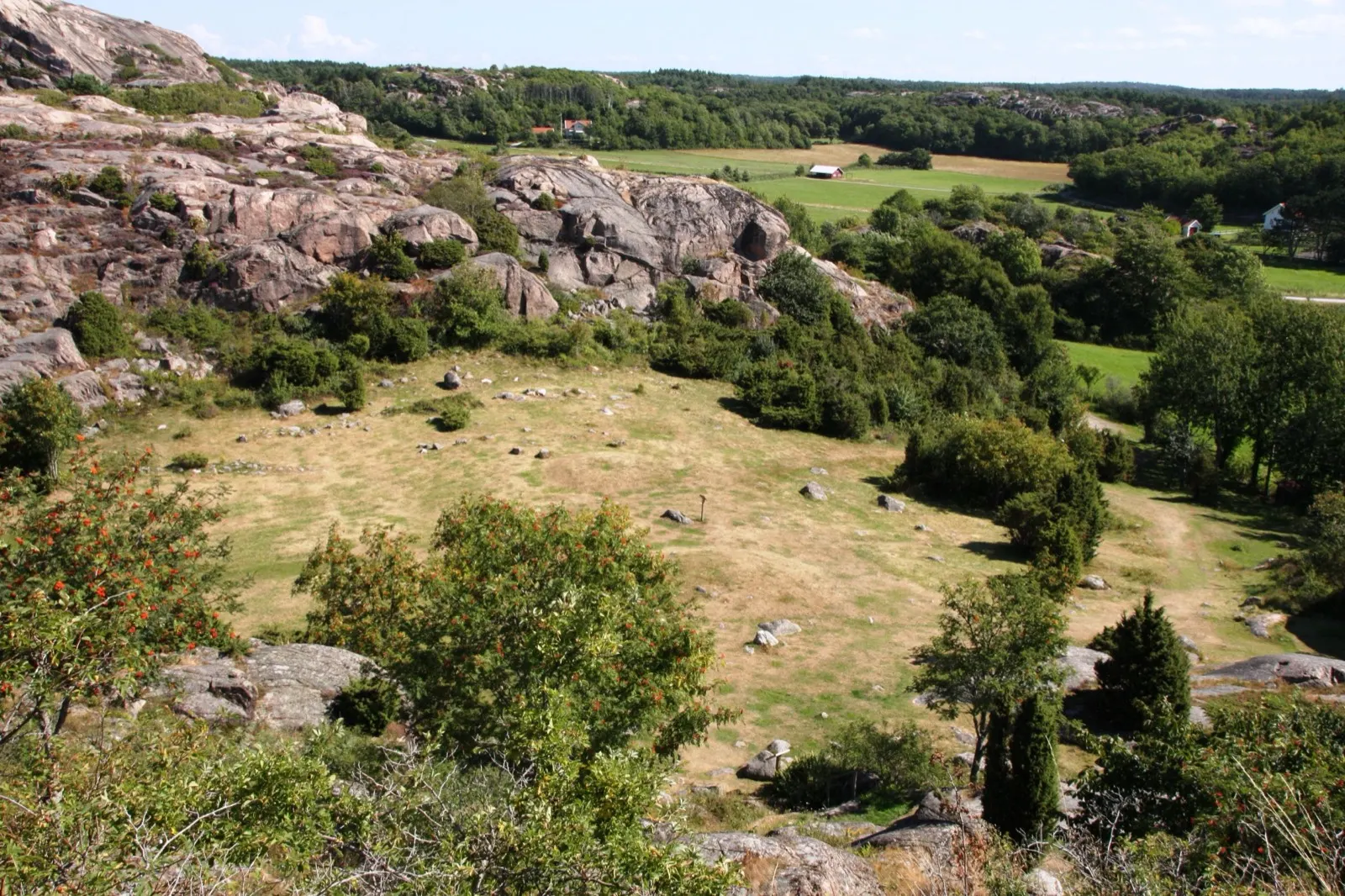 A grassy hollow ringed by bare granite outcrops at Hoge Slott, with farmland and wooded hills in the distance