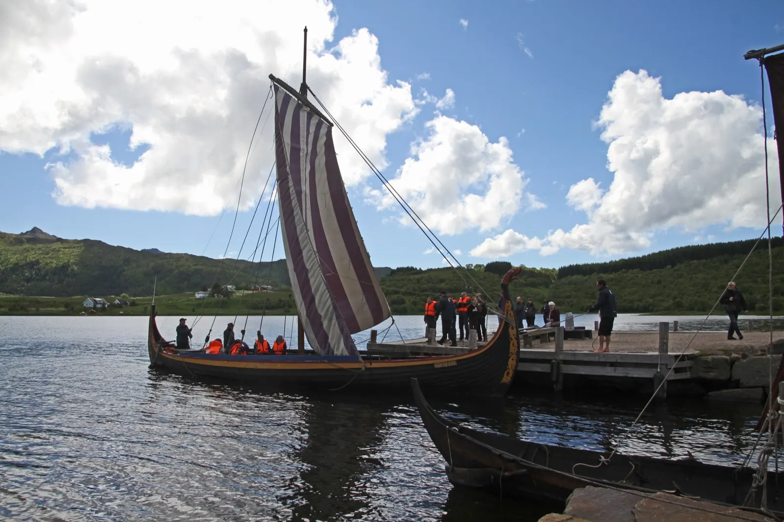 A Viking ship replica with a striped red and white sail and crew in orange life vests leaving a wooden jetty, green hills and a cloudy sky behind