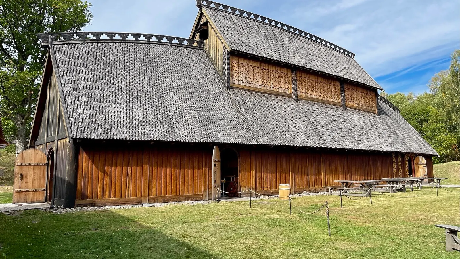 The Gildehallen at Borre, a long timber hall with a shingled roof, carved gable panels and picnic tables on the grass in front, under a blue summer sky