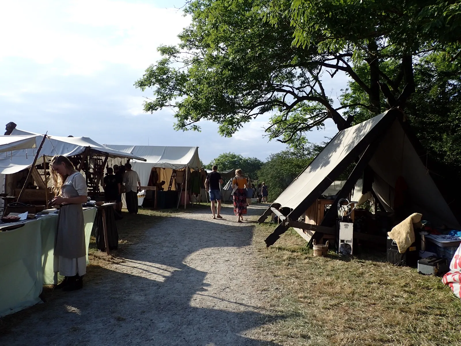 A gravel path through the market camp at Moesgaard with canvas stalls, a costumed woman at a table and a tent under a large tree in evening light