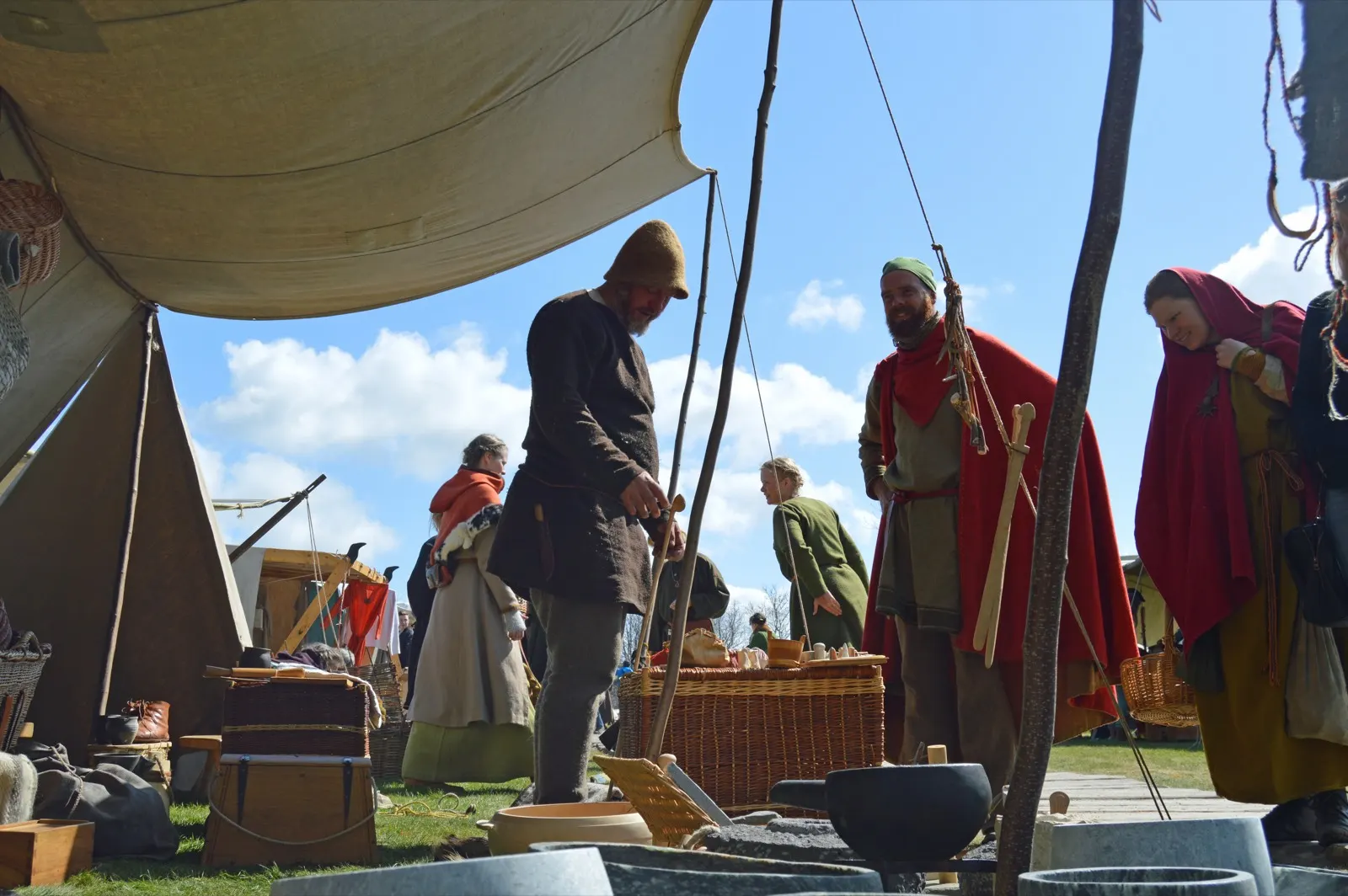 Costumed traders under a canvas awning at Ribe's international Viking market, a man in a straw hat leaning on a staff, a woman in a red hood and baskets of goods