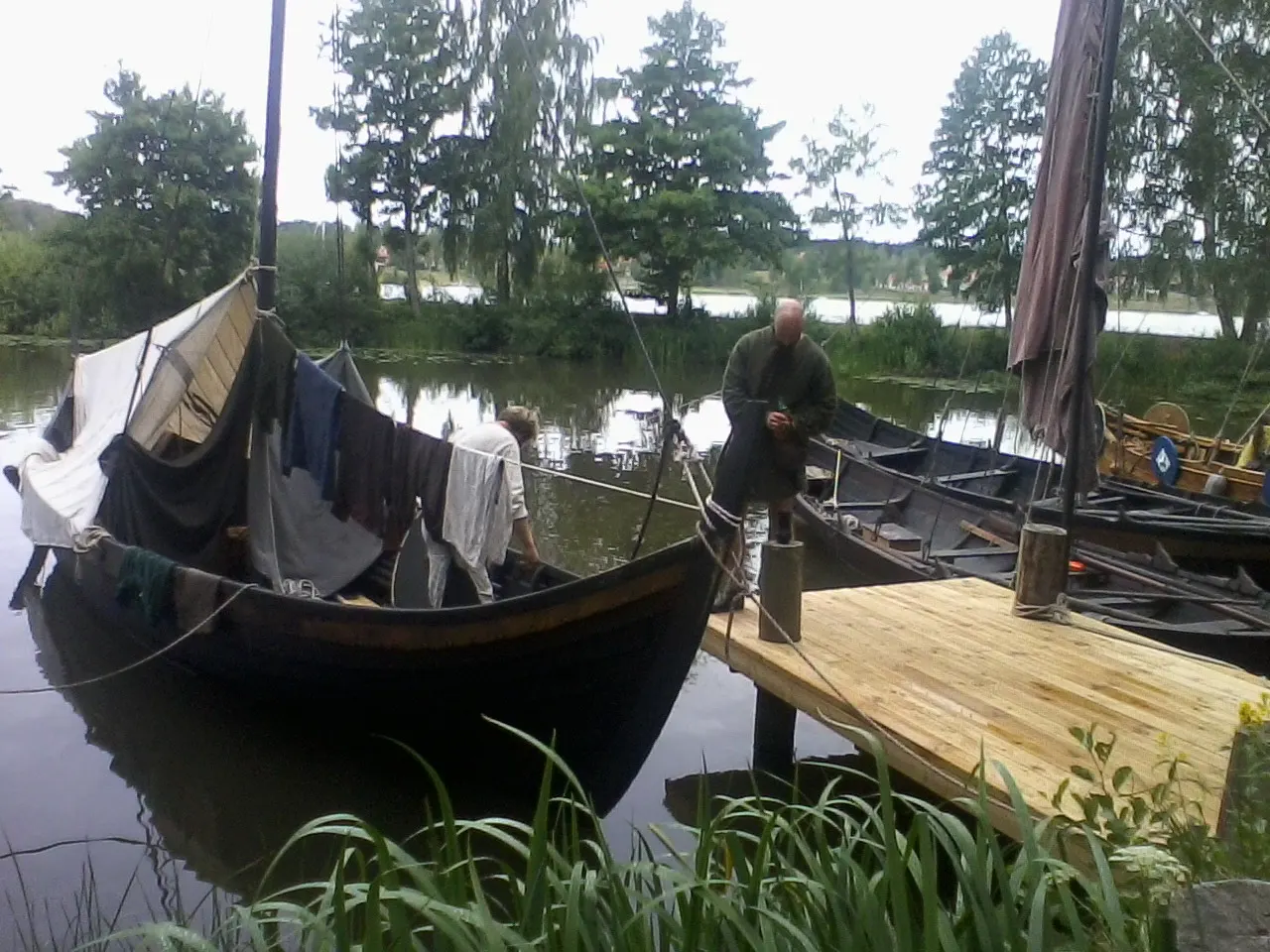 Two Viking-style wooden boats with a sail rigged as a canopy moored at a small jetty on the Mälaren shore, a man in a grey tunic standing on the pier