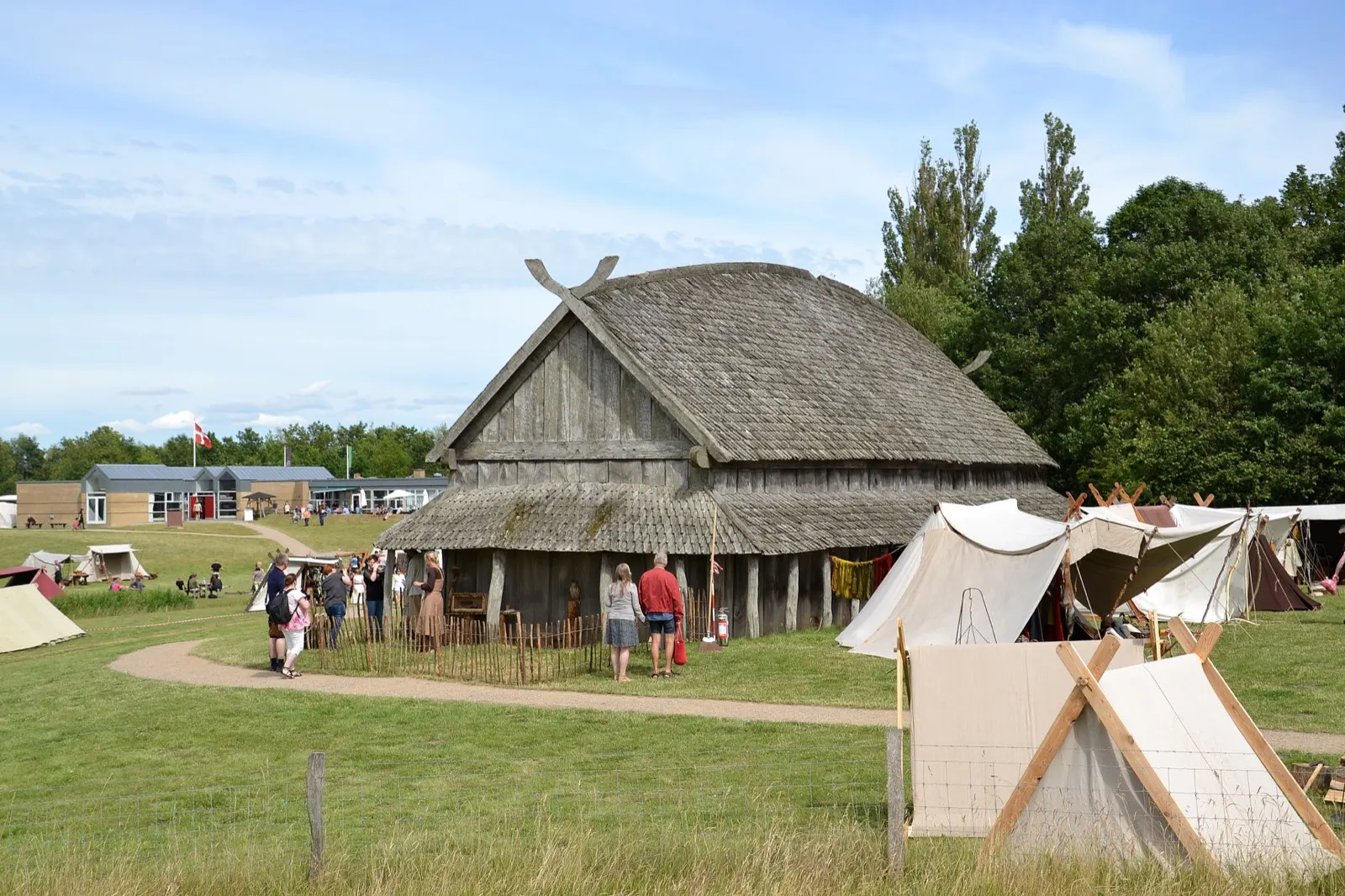 The reconstructed Trelleborg longhouse with a shingled roof and crossed gable beams, festival tents pitched in front and visitors at the fence