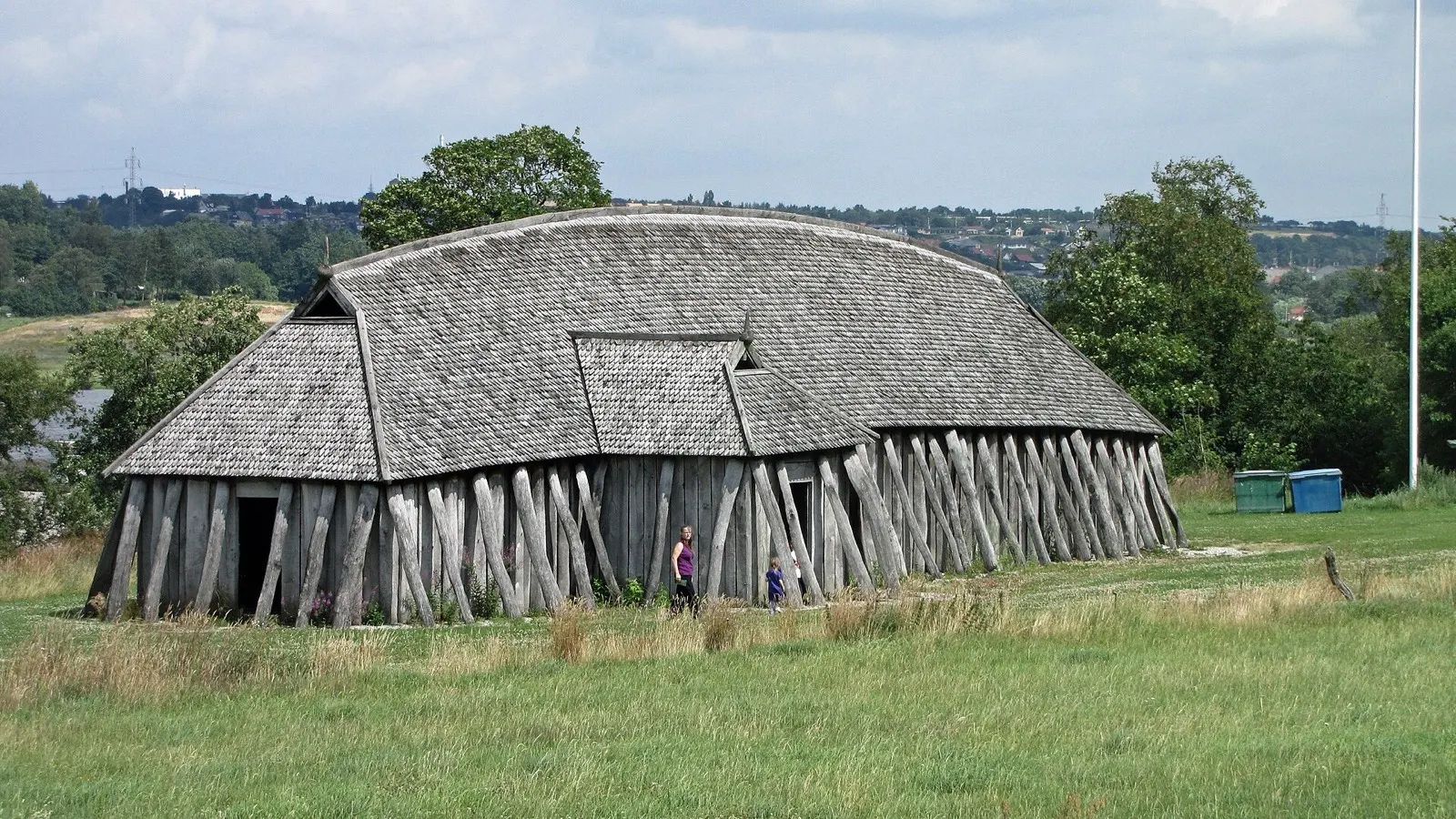The reconstructed Viking longhouse at Fyrkat, a long curved roof of wooden shingles over walls of sloping timber posts, on a grassy rise